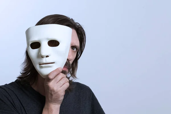 Uzun saçlı, bıyıklı bir adam yüzüne yakın beyaz bir maske tutuyor ve tek gözüyle dışarı bakıyor. Kimlik gizleme, gözetleme, verileri hackleme kavramı. Kopyala lütfen. Dikkat dolandırıcılar..