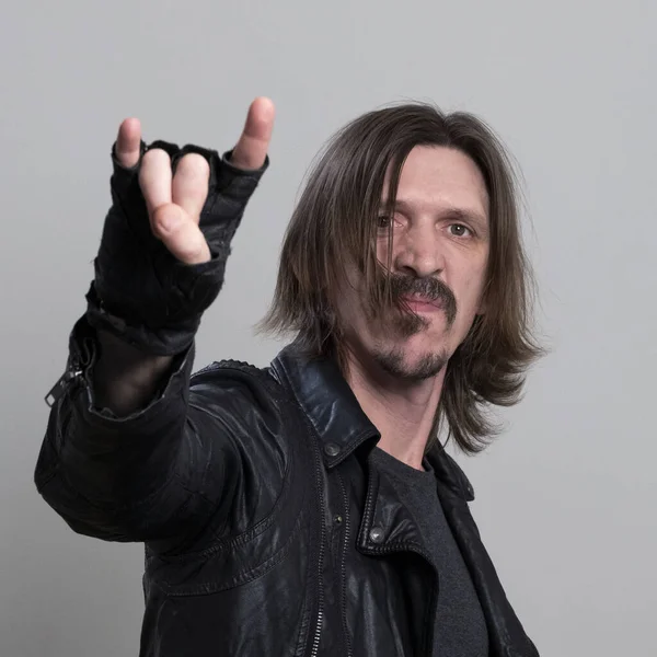 Uomo dai capelli lunghi in giacca di pelle nera e guanti fa un gesto rock su uno sfondo bianco. Concetto musicale heavy metal. L'immagine di un fan della roccia. Telaio quadrato . — Foto Stock