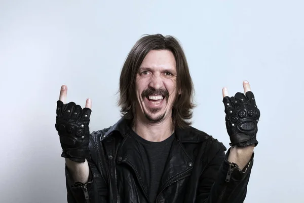 Un homme aux cheveux longs souriant vêtu d'une veste et de gants en cuir noir fait un geste rock sur un fond blanc. Concept de fan de rock ou de heavy metal . — Photo