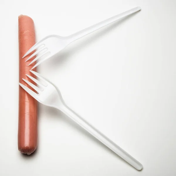 A sausage and two forks on a white background look like the letter K . The concept of fast food, hunger or lack of food. Environmental issue and ban on plastic dishes, poor student or senior citizen. — Stock Photo, Image