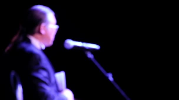 Homem de cabelos longos abstrato na frente de um microfone em um fundo escuro. Desfocado. Discurso de um empresário ou formador de nacionalidade asiática no palco em frente a uma audiência. Espaço de cópia para texto ou logotipo . — Vídeo de Stock