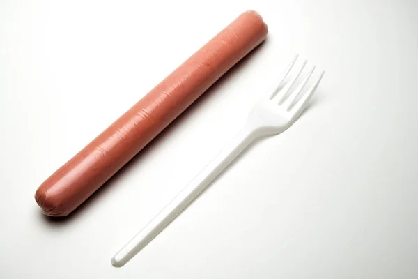 Sausage next to a fork on a white background. Copy space. The concept of fast food, GMOs, hunger or lack of food. Environmental issue and the ban on plastic dishes. — Stock Photo, Image