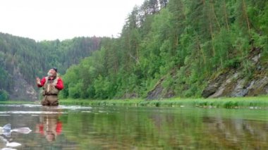 Kırmızı elbiseli bir balıkçı oltayla suya yem atıyor. Sinek avı. Dağ nehrinde spor ve eğlence. Genel plan. Doğa, bitki ve hayvan..