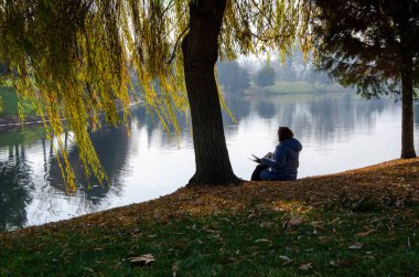 Kadın nehir kenarında bir dergi okuyor. 