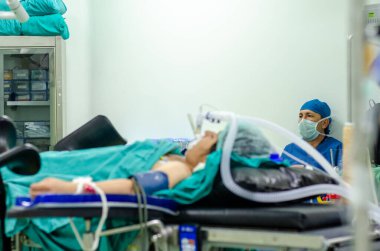 Ameliyathanede erkek cerrah oturuyor. Ön planda trakeal entübasyon tüpü olan bir hasta var.