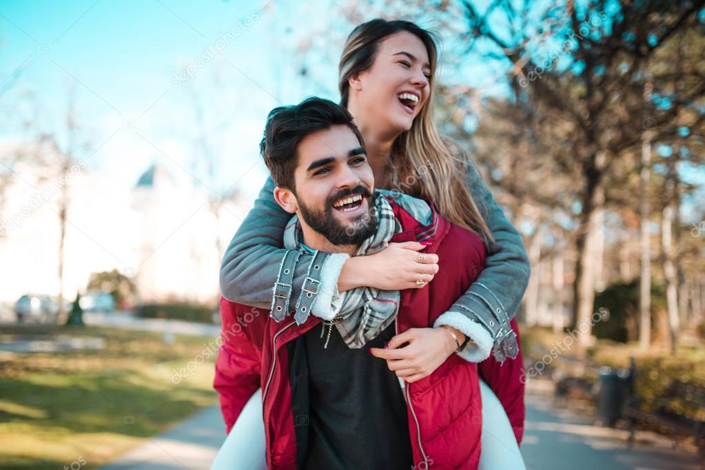 Couple in love having fun in the park. Man giving piggyback to woman. Young adults girlfriend and boyfriend hugging happy.