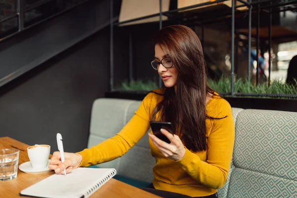 Woman writer using phone while drinking coffee and writing notes.