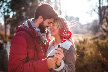 Güzel genç çift birlikte parkta öpüşme ve kalp şeklinde bir lolipop tutan, tadını seviyorum.