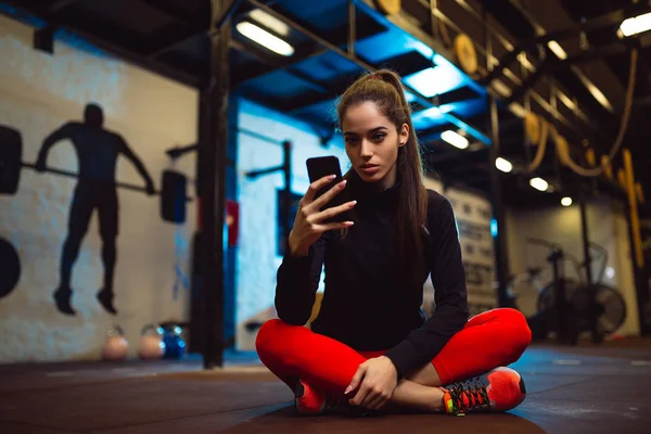 华丽的运动女孩坐在地板上的健身房和使用手机 — 图库照片