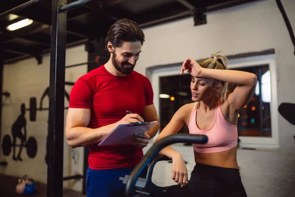 Exhausted young blonde girl finished her workout on bike in gym and talking with personal coach. Handsome personal coach writing results.