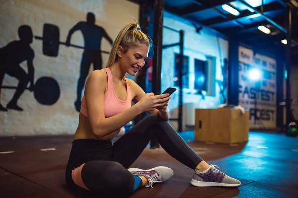 微笑的女孩坐在地板上 在健身房使用智能手机 — 图库照片