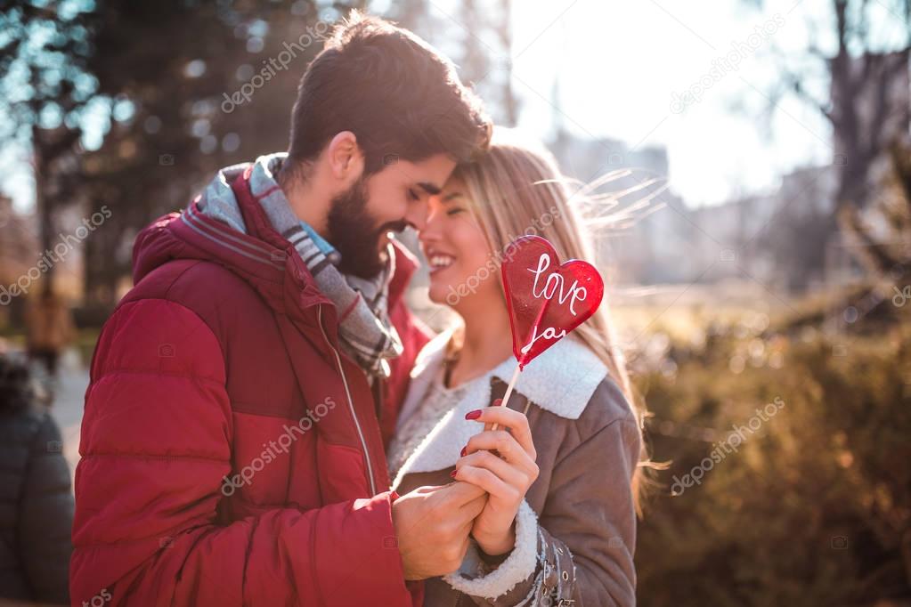 Beautiful young couple in love enjoying together in the park, kissing and holding together a lollipop in heart shape.