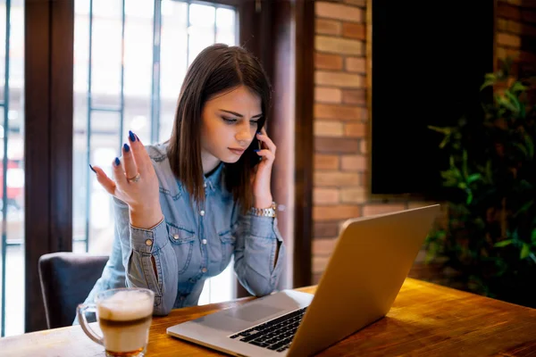 年轻的不满困惑的商业妇女坐在室内 在咖啡馆使用笔记本电脑和交谈的手机 — 图库照片