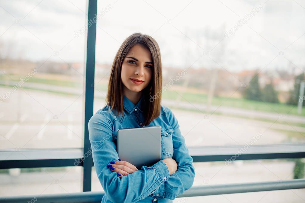 Portrait of confident smiling businesswoman holding tablet. Girl standing with arms crossed in office.