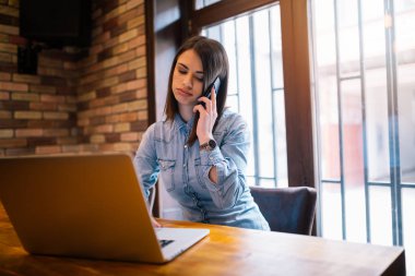 Hareket eden telefon ile tahta masada otururken bir gülümseme ile muhteşem kadın konuşan dizüstü bilgisayar, genç öğrenci veya iş ara, iş kadını olan açık net-kitap modern kahve dükkanı iç. Yorgun kız işitme kötü haber.