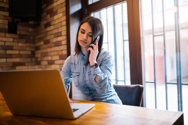 Hareket eden telefon ile tahta masada otururken bir gülümseme ile muhteşem kadın konuşan dizüstü bilgisayar, genç öğrenci veya iş ara, iş kadını olan açık net-kitap modern kahve dükkanı iç. Yorgun kız işitme kötü haber.