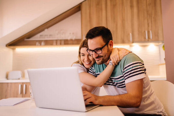 Young man with glasses is working on his laptop while his girlfriend is hugging him