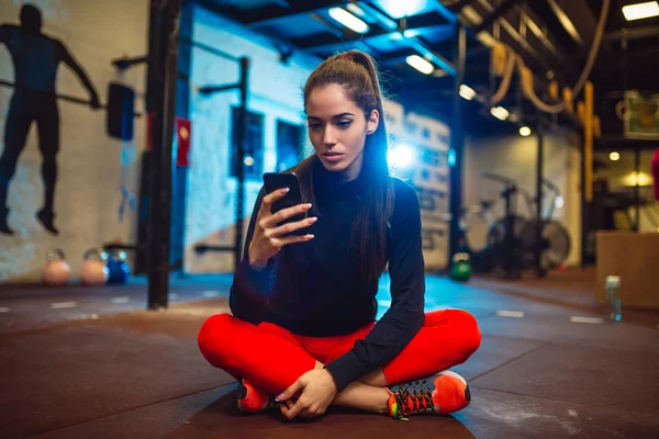 迷人的年轻黑发人坐在地板上健身房和使用她的智能手机 — 图库照片