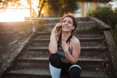 Genç kadın merdivenlerde açık havada otururken cep telefonuyla spor giyim