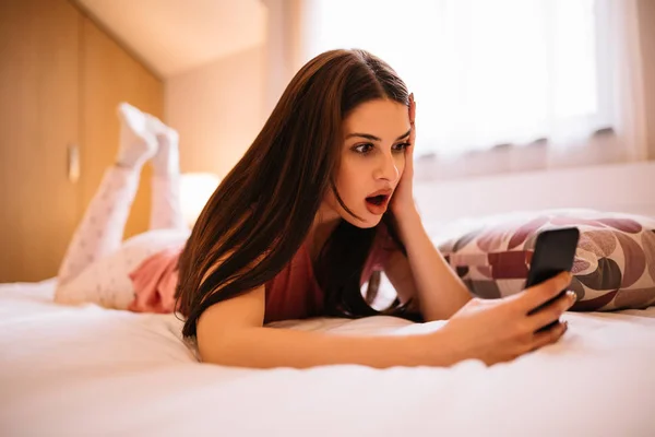 Здивована Молода Жінка Дивиться Телефон Лежачи Ліжку Вдома — стокове фото
