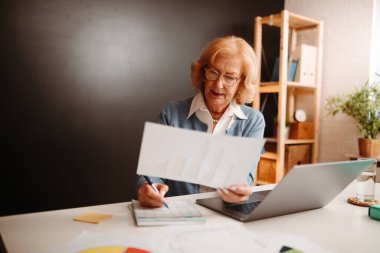 İş kadınının baskılı grafik tablosundan okunan verileri yazdığı yakın bir fotoğraf. Ofiste dizüstü bilgisayarın önünde oturuyor..