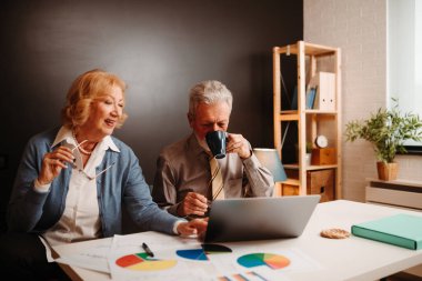 Ofisteki iş ortaklarının laptopun önünde otururken çekilmiş bir fotoğrafı. Gömlek ve kravat takan yaşlı adam kahve içiyor..