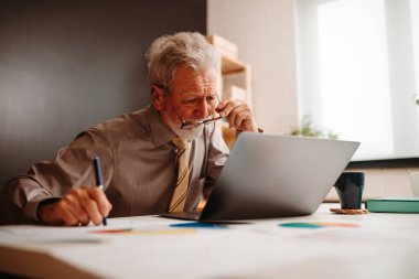 Ofiste dizüstü bilgisayara bakan yaşlı işadamının portresi. Bir fincan kahve dizüstü bilgisayarın yanında duruyor..