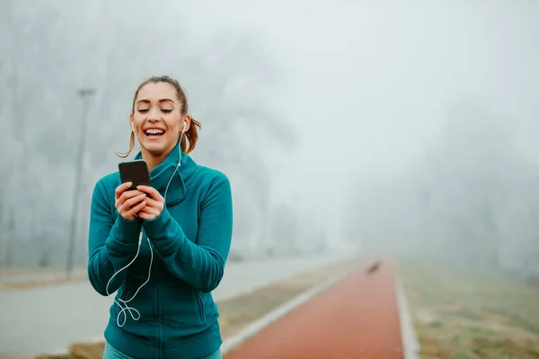 Training session could be fun. Young girl is sending text messages from her smartphone while jogging outside on foggy day.