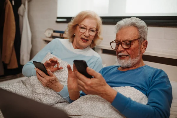 Portrait of elderly couple lying in bed and browsing online newspapers on their smartphones. Elderly couple using modern technology while lying in bed.