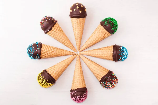Chocolate cake pops in waffle cone for ice cream in assortment, located in a circle, decorated with confectionery sprinkles on white wood table. Picture for a menu and confectionery catalog. Top view.