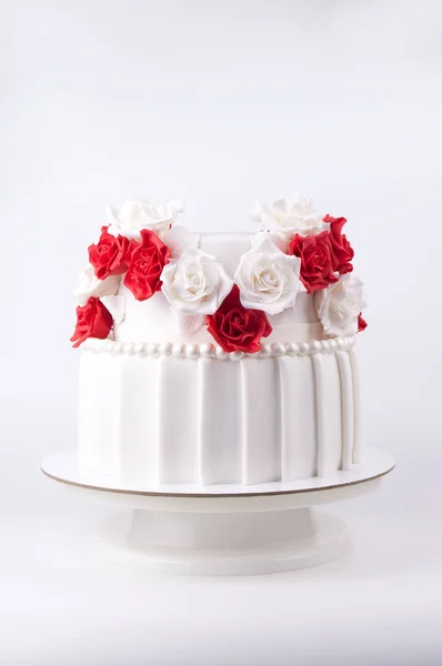 Two-tiered wedding cake decorated with red and white flowers from mastic on a white wooden table.