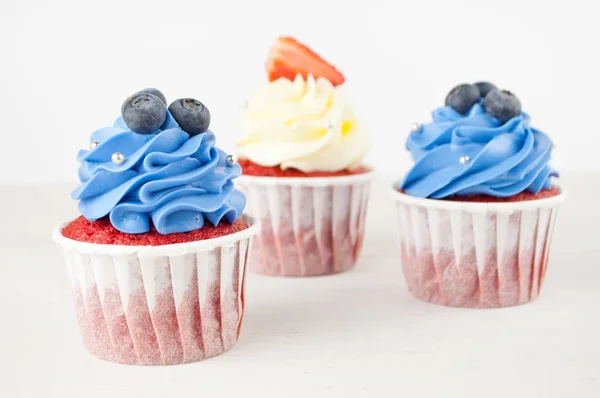 Cupcakes red velvet with blue and white whipped cream decorated with blueberry, strawberry on white wood table. Picture for a menu or a confectionery catalog.