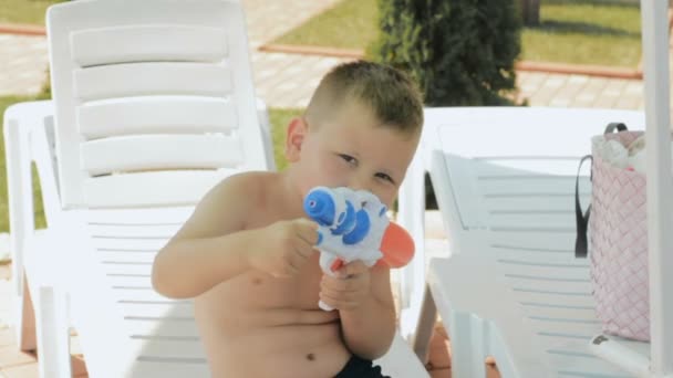 Liten pojke med en vattenpistol som sitter på en solstol och leende — Stockvideo