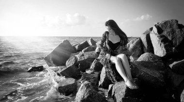 Yazın kumsalda genç bir kadın deniz kenarındaki taşlarda oturur.