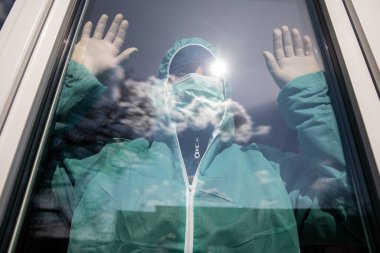 Maskeli bir adam kendini n-cov19 Coronavirus salgınından korumak için kapalı pencereden dışarı bakar.
