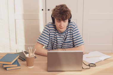 Kulaklıklı hoş bir genç çocuk dizüstü bilgisayar kullanır ve odasındaki masada öğrenir. O bir kurşun kalemle not alır. Salgın koronavirüs yüzünden uzaktan öğrenme