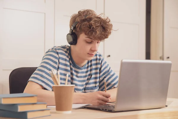 ヘッドフォンの素敵な10代の少年は ラップトップを使用して 彼の部屋の机で学びます 彼は鉛筆でメモを取る 伝染病のコロナウイルスのための距離学習 — ストック写真