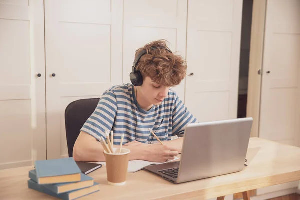 ヘッドフォンの素敵な10代の少年は ラップトップを使用して 彼の部屋の机で学びます 彼は鉛筆でメモを取る 伝染病のコロナウイルスのための距離学習 — ストック写真