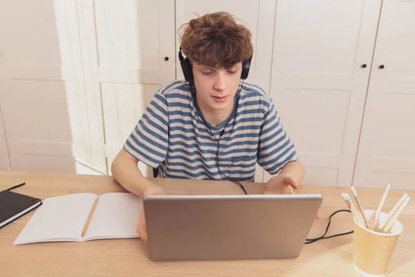 A nice teenage boy in headphones uses the laptop and learns at the desk in his room. He takes notes with a pencil.