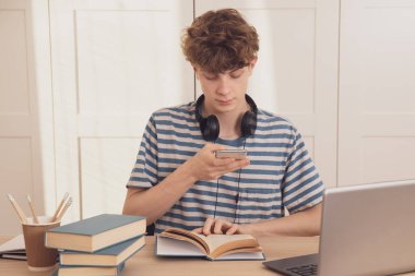 Kulaklıklı genç, notlarının fotoğraflarını telefonla çeker ve onları sınıf arkadaşlarına gönderir. Laptop, telefon ve tablet kullanarak öğrenir. Salgın koronavirüs yüzünden uzaktan öğrenme.