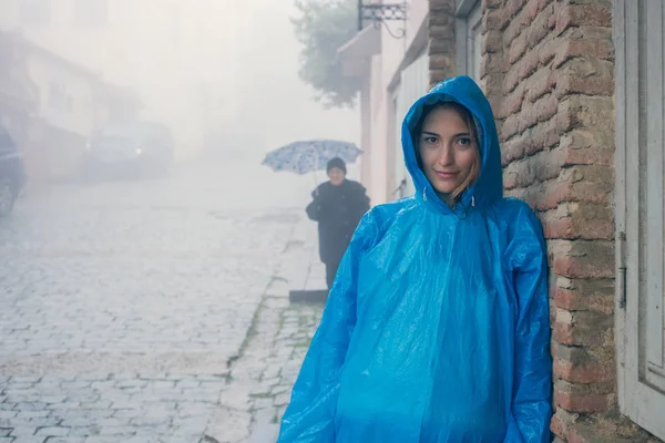 Young fashion woman in blue raincoat standing in mist outdoors. Brick wall of the old house. Pavers. A drizzle of rain. Gloomy atmosphere