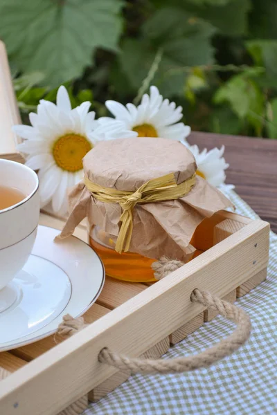 Zomertijd thee met kamille thee en honing in een glazen pot, bloemen en natuurlijke houten dienblad, met groene textiel servet in een kooi aan de houten bruine tafel Rechtenvrije Stockfoto's