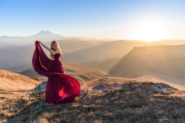 Gün batımında, dağların arkasında, kırmızı uzun elbiseli genç ve güzel bir kız. Sevgililer Günü. Metin için boşluk. Kafkas Dağları