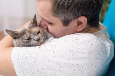 Bir adam bir kediye sarılır. Evdeki bir odada kanepede kediye sarılıyor. Ev eşyaları, mezosalin. Evcil hayvanlarla evde kal. Bir adamın ellerinde mutlu bir kedi yavrusu.