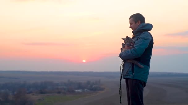 日没時に猫を連れた男ペットと春の散歩 猫は男の手に座っている 地平線の夕焼け空 — ストック動画