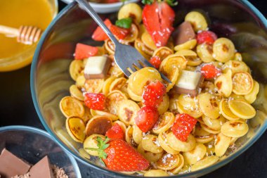 Mini krep gevreği kasesi taze çilekli muz çikolatası, hindistan cevizi talaşı ve küçük tabakta bal. Yukarıdan manzara. Kendini soyutlama eğilimi.