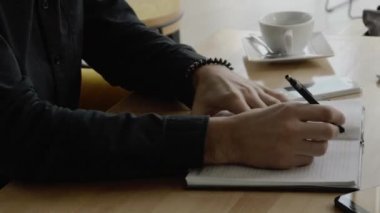 Kafenin ahşap masasında oturan, elinde kalem ve defter olan bir adamın elleri..