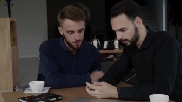 Deux hommes d'affaires joyeux discutant de quelque chose et souriant tout en étant assis au café avec un ordinateur portable et des téléphones. Deux jeunes collègues qui ont réussi à discuter des moments d'organisation par table dans un café . — Video