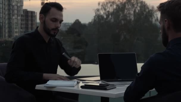 Dos hombres de negocios discutiendo algo y sonriendo mientras se sienta al aire libre con un ordenador portátil y teléfonos en el tiempo de la noche. Dos jóvenes colegas exitosos discutiendo momentos organizacionales . — Vídeos de Stock