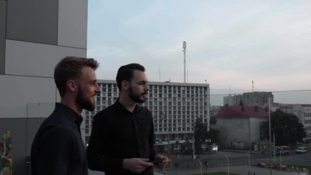 Jovem empresário colegas de trabalho sorrindo e discutindo algo juntos, enquanto em pé ao ar livre em um piso alto ao pôr do sol hora da noite. Parceria empresarial ou diálogo entre colegas . — Vídeo de Stock